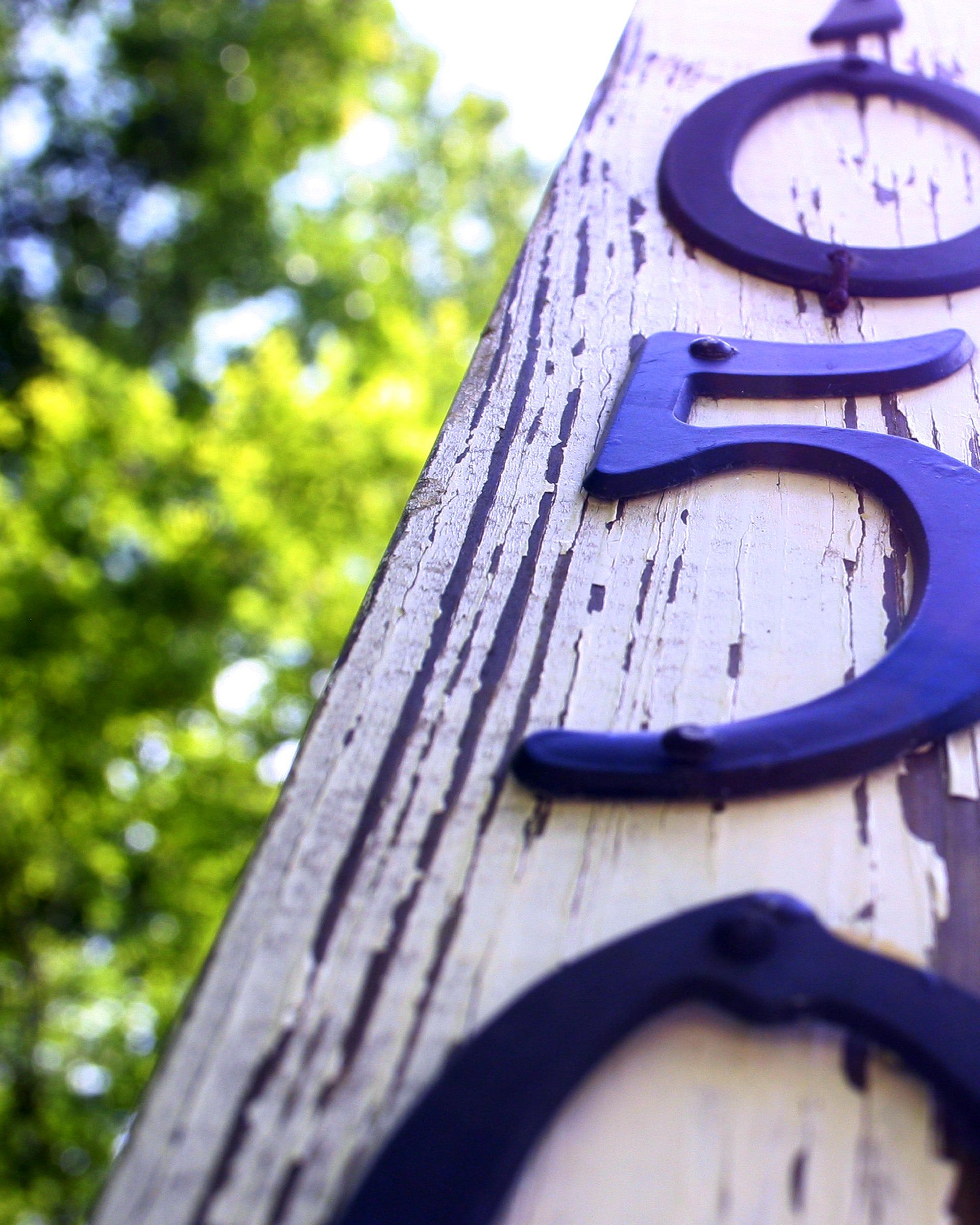 Blue-violet house numbers on a weathered white post, with green trees behind